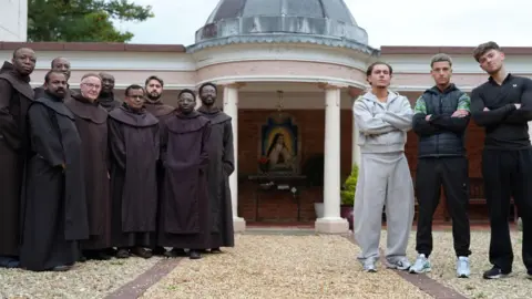Channel 5/Orchard Studio Three young men standing with their arms crossed with a group of traditional friars standing beside them in a courtyard.