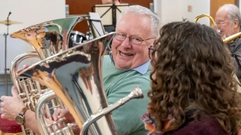 A man and woman playing a brass musical instrument. They are sitting down. The woman has her back to the camera whereas the man, who looks jovial, is looking towards the camera.