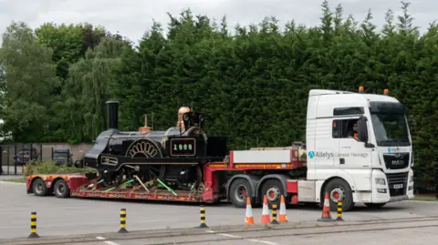 The Board of Trustees of the Science Museum Group A black locomotive is on the back of a white and red low-loader vehicle on tarmac. There are red and white and black and yellow cones on the tarmac as well.