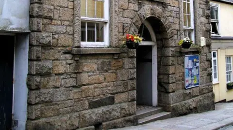 A brown brick building with a large arched door. There are large windows either side of the door. There are flowers in baskets. 