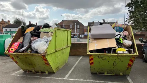 Boston Borough Council Two lime green skips full of rubbish in a car park side by side. One has a mattress and a large piece of carboard, as well as a bike frame, while another has plenty of bags full of rubbish