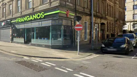A city street with stone shopfronts and parked cars. 