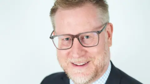 NSFT Jason Hollidge is smiling at the camera, it is a shot of juts hos head and you can just see the top of his shoulders. He is wearing a blue suit and light blue and white checked shirt. The background is white. Hollidge has a beard and is wearing glasses.