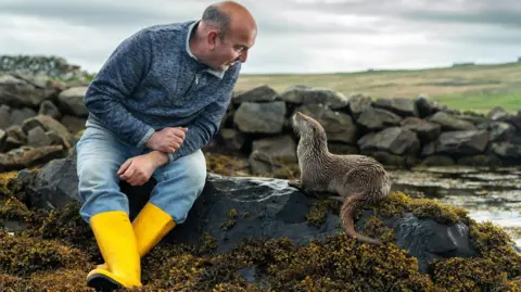 National Geographic Billy Mail is sitting next to Molly the otter on rocks near the sea