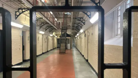 A general view picture of the ground floor of Reading prison, with cells on the left hand side of it. There is a black staircase up to other floors in the centre of the ground floor.
