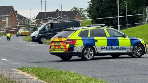 A police car parked across a road behind it there is police tape and a person in hi-vis clothing.