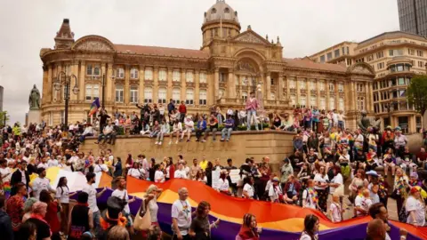 John Bray Hundreds of people gathered in front of a stone city council building. They are taking part in or watching the parade at Birmingham Pride. People are walking through the middle of the street with a purple and orange flag.