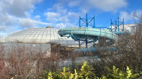 A dome and a blue curvy leisure centre slide are seen from behind a metal fence and shrubs.