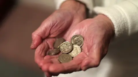 A close-up of an older person's hands, holding pound coins in the palm of their hands. They are wearing a white jumper