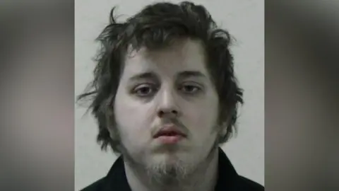 Mugshot of Dorritt. He has long scruffy brown hair, a faint moustache and wispy fair hair on his chin.
