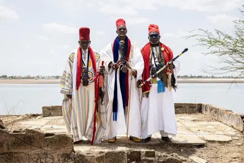 PATRICK MEINHARDT / AFP / GETTY IMAGES Three men dressed in boubous, tall red hats and carrying batons, pose for a photograph next to a body of water.
