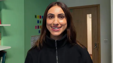 Anika Kaul, a woman with long dark hair, wearing a black zipped top, smiles at the camera