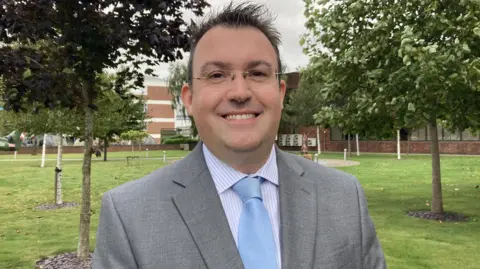 Adam Clarke, wearing a grey suit and blue tie, stood in the grounds of Leonardo, smiling at the camera.