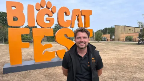 Cameron Whitnall standing in front of a big orange sign which reads 'Big Cat Fest' with the newly completed Small Cat Rescue Centre to the right
