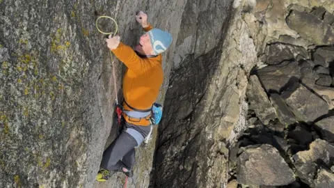 Rob Matheson Rob Matheson is climbing a practically vertical rock face with very few parts poking out to provide any sort of hold. His right hand appears to be hanging on by no more than his fingertips. There are large grey boulders below him. He is wearing an orange fleece, a blue hat and black trousers. With his left hand he is reaching for a thin climbing hold on the rock.