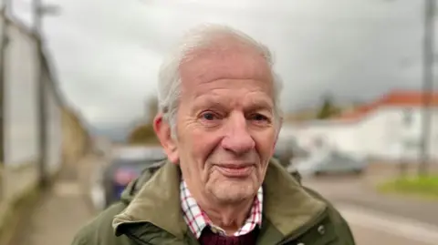 Martin Giles/BBC Gerald Kelly is standing on a paved street with buildings and parked cars in the background. He is wearing a green jacket over a red-and-white checked shirt. The sky appears overcast.