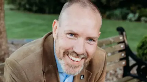 AMORÉ DU PLESSIS PHOTOGRAPHY Graham smiles for the camera. He is sat on a bench, wearing a brown corduroy suit and shirt. He has a shaven head and stubble.