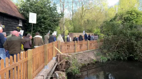 Crowds of people holding banners and walking alongside the edge of a river which has fencing along it.