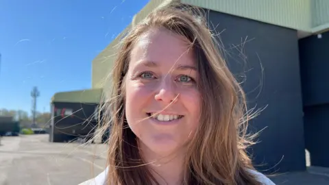 A woman with medium-length brown hair which is blowing in the wind. She has blue eyes. She is smiling. Behind her is the LNER Stadium.