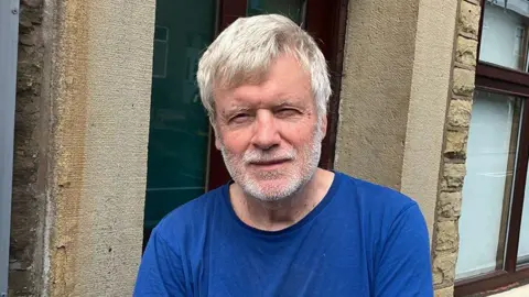 David Daly with grey hair and beard wearing a blue t-shirt stands in front of a house.