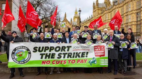 A small crowd of people with dark clothing standing outdoors with a large green and white banner which reads support the strike, not cuts to pay