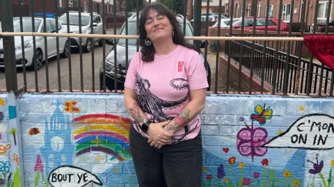 A lady with brown hair, wearing a pink top stands in front of a bright, blue wall. Signs on the wall say "bout ye" and "c'mon on in"