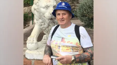 Cumbria Police A picture of Stanley Smith. He is smiling and is wearing a blue bucket hat with the words Sergio Tacchini written on in white lettering. He appears as though he is on holiday in a park. A stone lion is behind him. He has a small stud earring and wears a white T-shirt. The straps of a black backpack can also be seen.