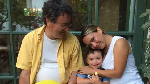 Oscar and Erica McLennan sit on either side of their wee boy Ian. Oscar is wearing a yellow shirt with white top undeneath, and is looking sideways at his wife and son. Ian is grinning cheerfully into the camera while being hugged by Erica, a woman with blonde hair and a white top on. They appear to be sitting outside, with glass panes behind them.