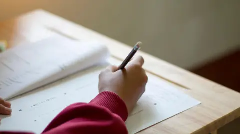 Getty Images A child's hand on a school desk. The child is holding a pencil and writing in what looks like an exam paper