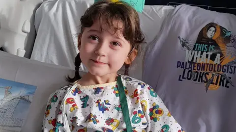 Oxford Hospitals Charity Sylvie during her treatment on Bellhouse Ward. She is looking away from the camera. She is wearing a white pijalams with various colourful characters on. Her hair is braided. She is lying on several cushions.