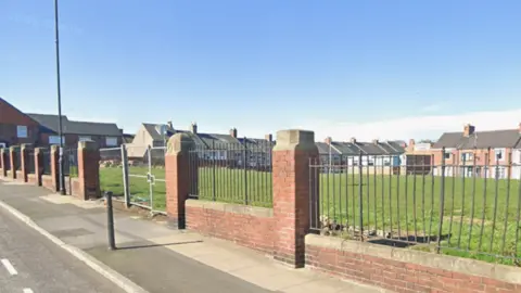 An open stretch of grassy land off Seaside Lane in Easington. The school was demolished several years ago. The site is surrounded by a brick wall and metal fence perimeter. 
