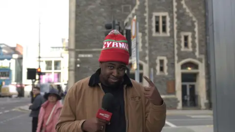 BBC reporter Terel Edmunds holding a BBC News branded mic and wearing a bobble hat which has the word Cymru misspelt as Cyrmu