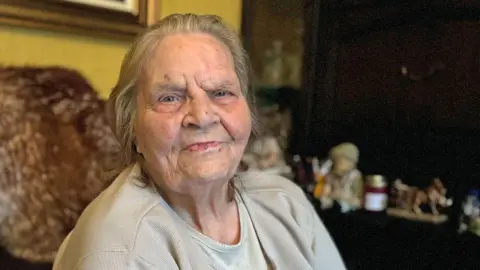 Annie Sweeney sitting on a chair in a living room.  She has straight, grey hair swept back from her face and is wearing a beige jumper and T-shirt. 