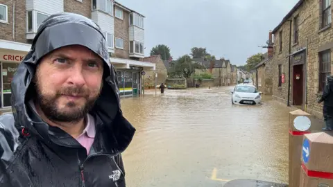 Tim Bearder Tim Bearder is wearing black coat with his hood up. Behind him is a street submerged in brown murky water.