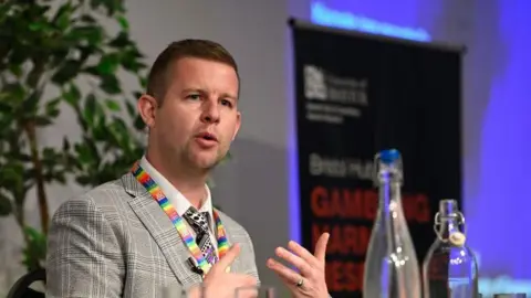 Dr Reece Bush-Evans Dr Reece Bush-Evans is pictured speaking at an event. He is wearing a checkered suit and a lanyard with the LGBT rainbow colours on the neck strap. 