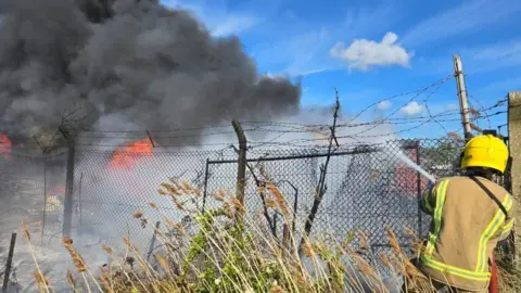 Firefighter on far right with water hose spraying though a metal fence onto flames.