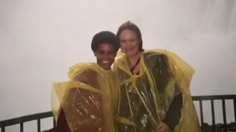 Christopher MacRae Christopher as a younger boy with his mum at Niagra Falls. They are wearing yellow ponchos and standing in front of a railing, with the water from the falls behind.