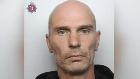 A mugshot of a man with a light beard and bald head. He is wearing a black hoodie. 