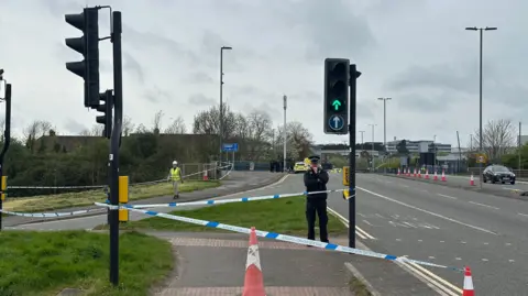 A police officer in uniform mans a cordon next to a busy main road. Lots of police vehicles can be seen parked in the background, and there is lots of blue and white police tape at the entrance of the road. 