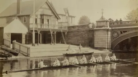 Christ's College A sepia photo shows a wooden-framed boathouse on the river bank and a bridge with people watching a rowing crew on the river below.