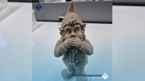 A cement-coloured gnome, made from MDMA, covers its mouth with its hands on a police desk