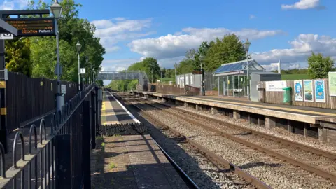 The picture is taken from a railway station, looking out onto the track as it stretches out into the distance. The sky is blue and the track is flanked by tall green trees. 