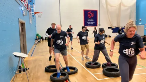 Men and women in matching t-shirts are running towards and between tyres laying flat on the floor of a sports centre