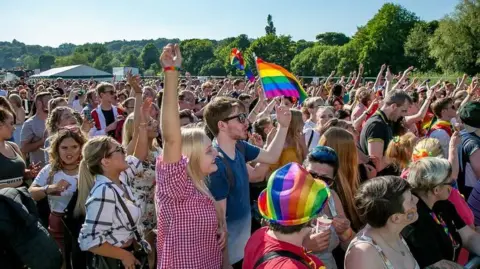 A crowd of several hundred people raise their hands in the air and wave rainbow flags during a Pride event in Durham.
