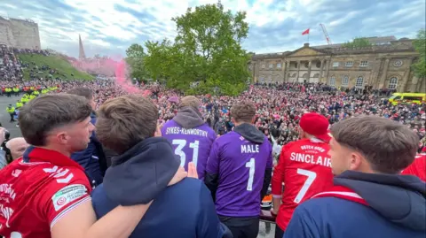 Oli Constable/BBC Players stand on the top deck of a bus and look across masses of supporters cheering them on.