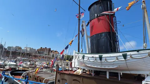 Stuart Maisner/BBC Dozens of little ships gathered in Ramsgate Harbour on a sunny day with a big tug in the foreground waving a union jack flag