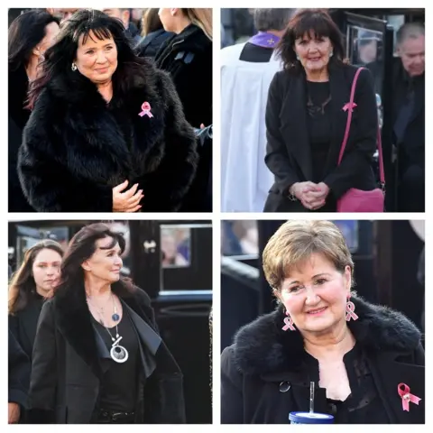 PA Media/Getty Images Coleen, Anne, Maureen and Denise attend their sister's funeral. Coleen wears a fluffy black coat and has dark hair. She has a pink ribbon on her coat. Anne has dark hair and carries a pink bag. Maureen has dark hair and has a necklace with the number 6 on it. Denise has shorter hair and has pink ribbons as earrings.