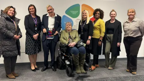 City of Wolverhampton Council A group of people stand in a line, with a woman in the middle sitting in a wheelchair. They all smile at the camera. A man in the middle holds a piece of paper saying 'finalist' on it.