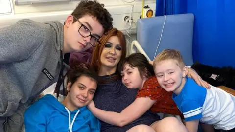 Moffat family Michelle-Anna Moffat has long red hair and is lying on a hospital bed. Her four children are crowding around her, smiling and hugging her.