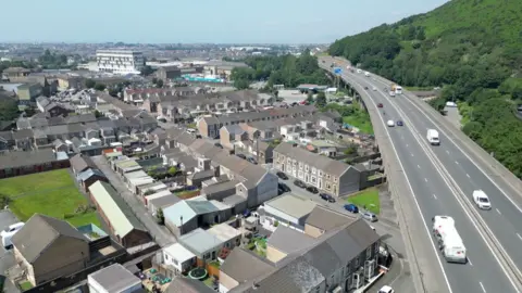 The M4 motorway can be seen on the right hand side of the image, on the left hand side is a tightly arranged group of houses. The motorway is raised above the land that the houses stand on, it has pillars that keep it elevated that overhangs some of the residential land, some of pillars that keep it elevated have been erected in the gardens of these houses. There are also more houses that stretch all the way to the horizon, as well as a massive cluster of trees on a hill. 